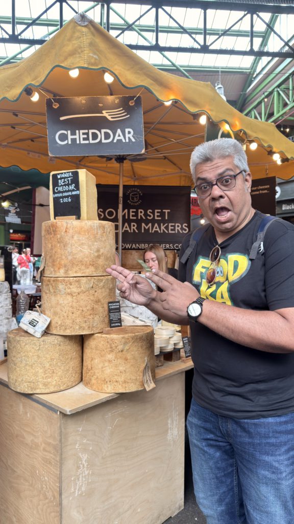 IMG_4357 - Social Nation Man posing at a cheese stall with stacked large cheddar wheels and a sign that reads'Cheddar' under a yellow canopy in a market.