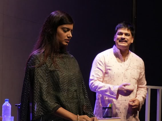 Two performers on a dimly lit stage: a woman in a dark patterned outfit looks down, while a man in a light pink shirt gestures beside her.