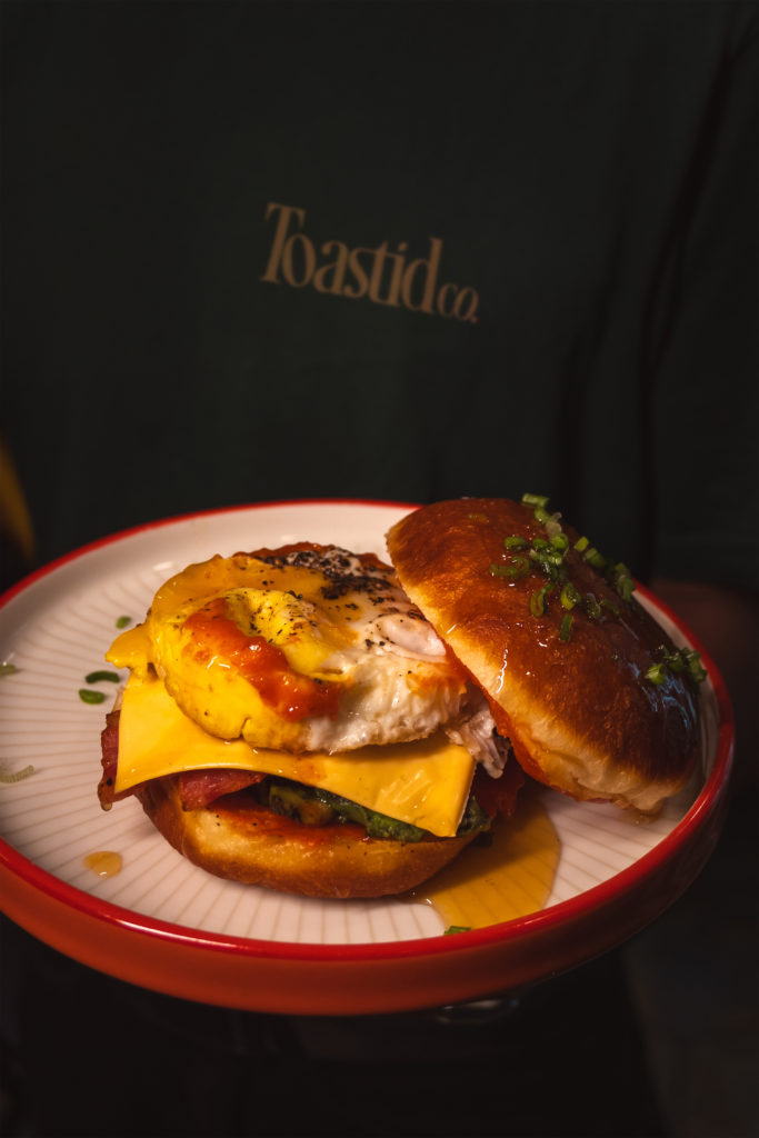 Breakfast Burg - Social Nation Plate with a bacon cheeseburger topped by a fried egg and melted cheddar, drizzled with sauce on a white plate with a red rim.