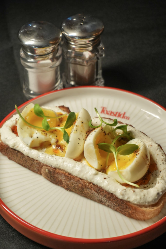 Chilli Egg Yogurt - Social Nation Open-faced toast topped with soft-boiled eggs, creamy sauce, and microgreens on a white plate with a red rim.