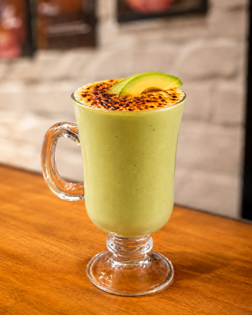 Avacado Avalanche - Social Nation Green matcha latte in a glass mug with a torch-charred top and an avocado slice garnish on a wooden table.