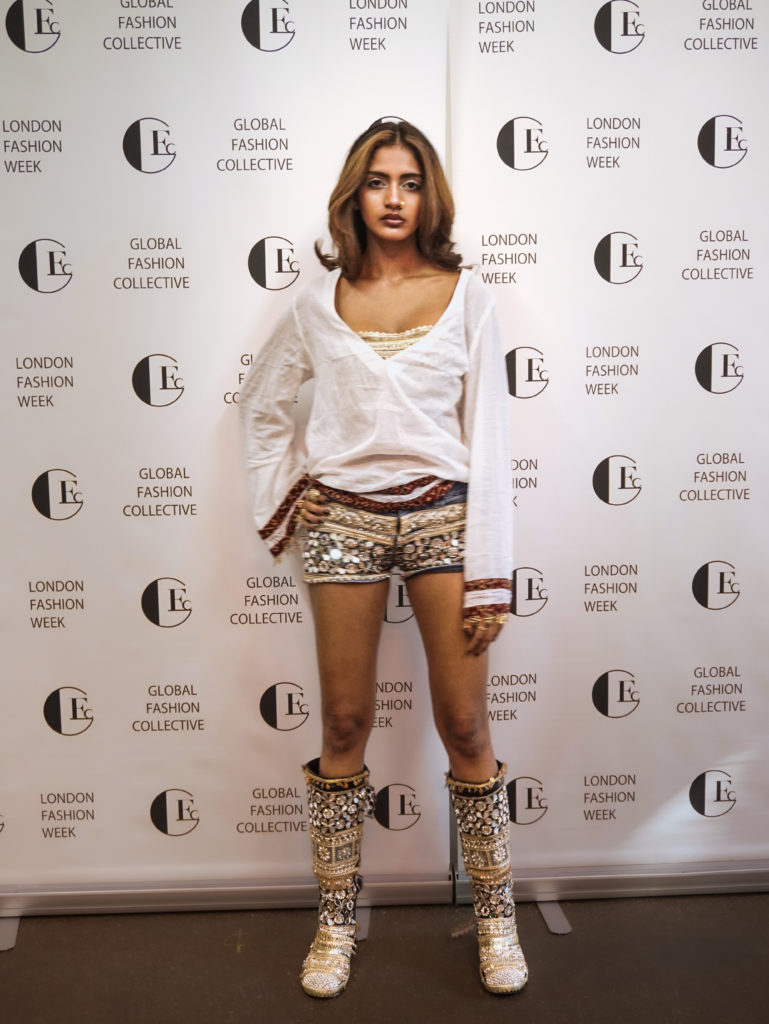 Ria Mehta - Social Nation Model posing in front of a London Fashion Week backdrop with Global Fashion Collective logos, wearing a white wrap top, patterned shorts, and embellished knee-high boots.
