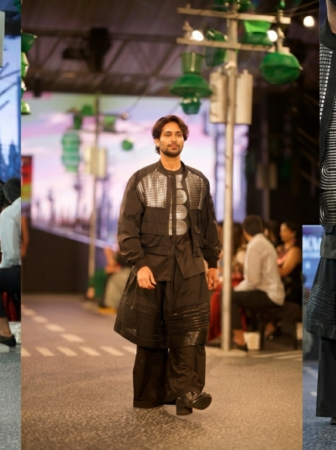 Male model on a runway wearing a black outfit with silver geometric panels and a wide, layered skirt-like coat.